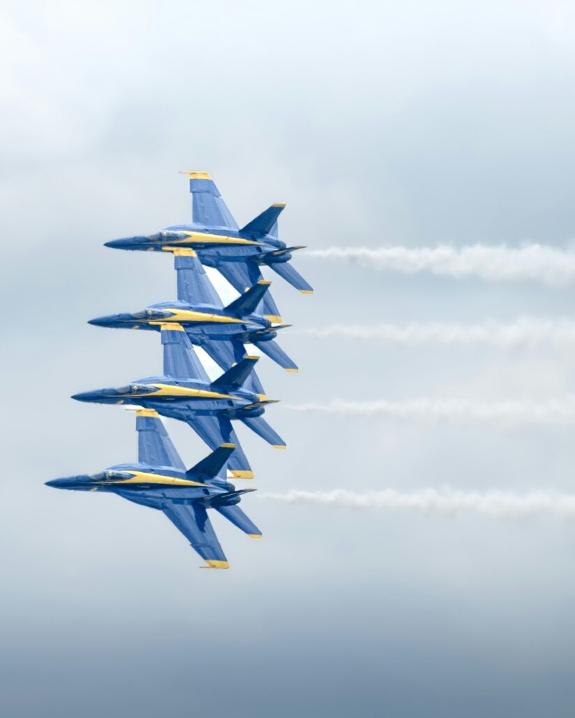 Four blue and yellow fighter jets fly in tight formation, leaving white vapor trails behind them as they soar through a cloudy sky. The aircraft are aligned closely, demonstrating precision and coordination in flight