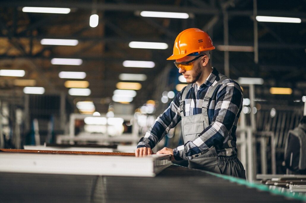 A factory worker wearing an orange hard hat, safety glasses, and a plaid shirt with overalls focuses on operating machinery or inspecting a product in an industrial setting. The background is filled with equipment and large structural elements, indicating a busy manufacturing environment.