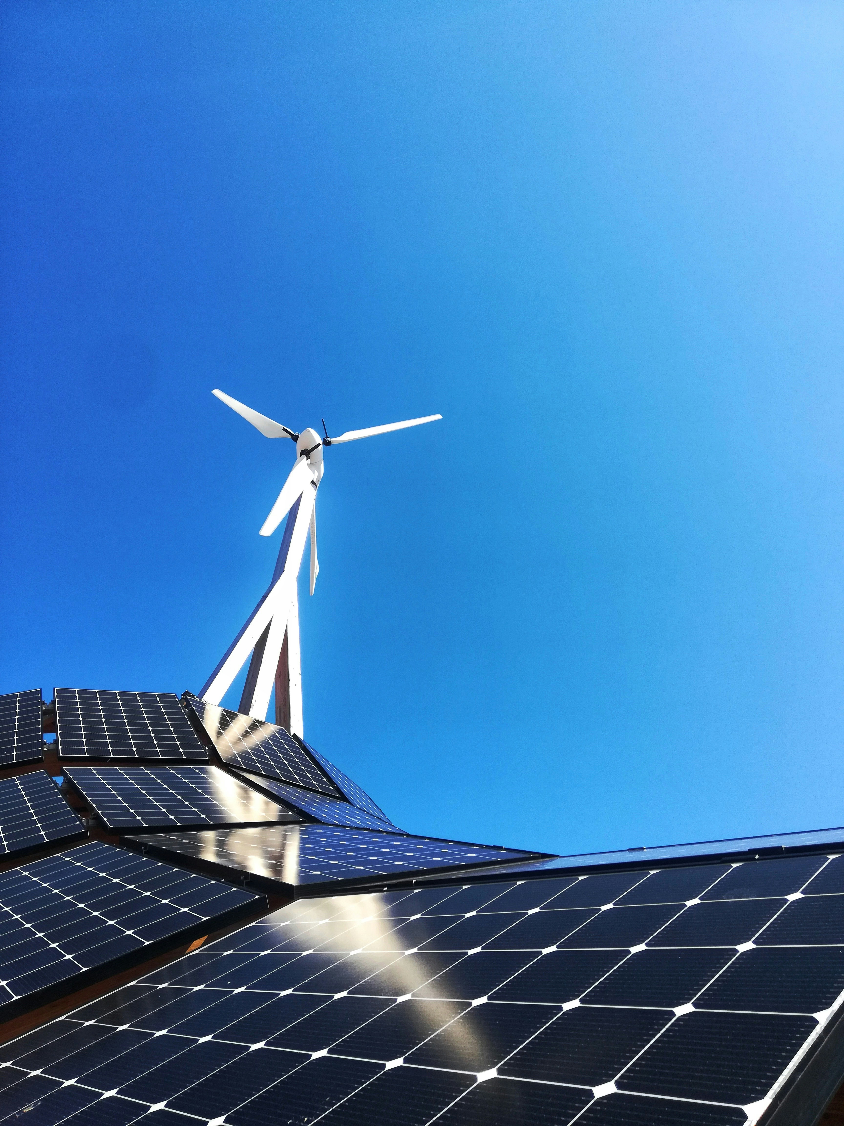 A close-up of solar panels with a wind turbine in the background, both set against a bright, clear blue sky. The image highlights the combination of renewable energy technologies, showcasing both solar power and wind energy.