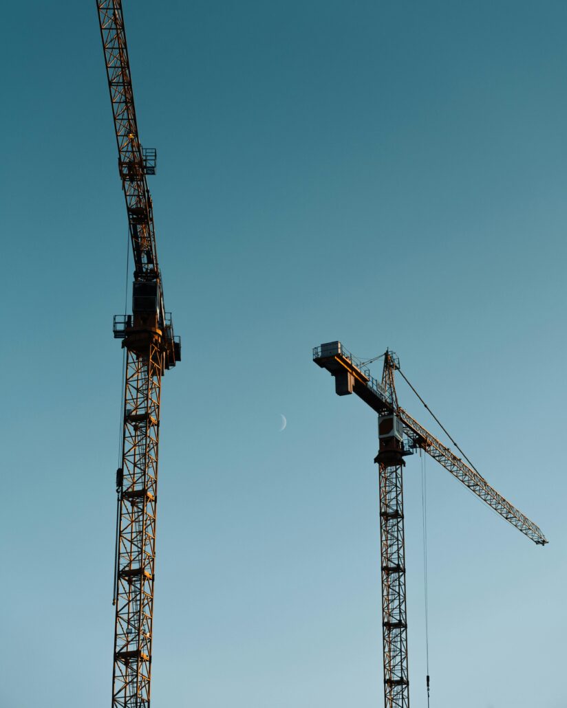 Two tall construction cranes stand against a clear evening sky with a crescent moon visible between them. The cranes are silhouetted against the soft blue background, emphasizing their height and structure.