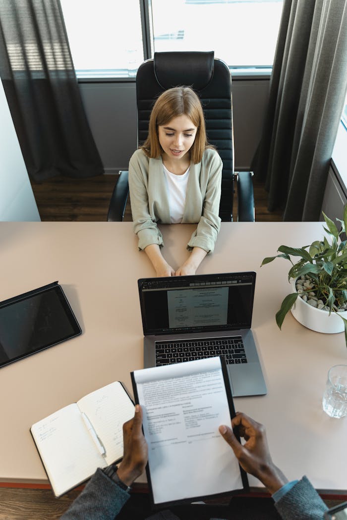 A top-down view of a job interview or meeting in a professional office. A woman sits across the desk with her hands folded while another individual, partially visible, reviews a resume or document. A laptop, notebook, and tablet are also on the desk, along with a potted plant and glass of water.