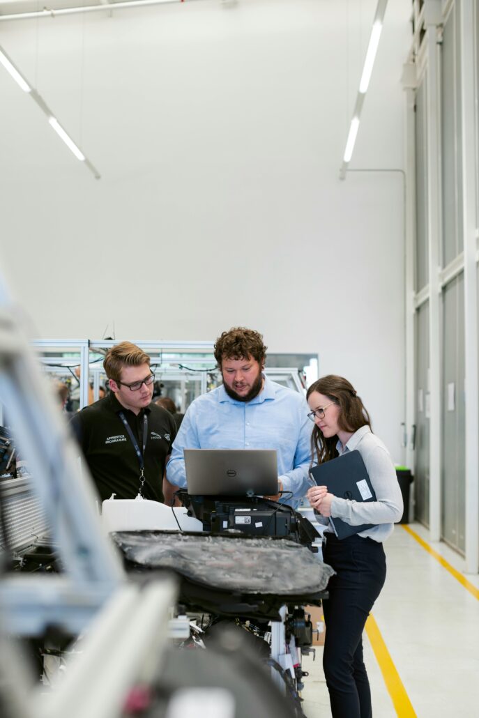 Three people in a modern manufacturing or lab setting gather around a laptop, engaged in discussion. One man in a blue shirt operates the laptop while a man in a black shirt and a woman holding a clipboard look on attentively. Machinery and equipment are visible in the background, and the space has a clean, industrial design with bright overhead lighting.