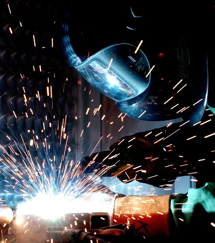 A welder wearing a protective helmet and gloves works on a metal project, producing bright sparks and intense light from the welding process. The image captures the dynamic energy of the welding operation, with flying sparks and a strong glow from the molten metal.